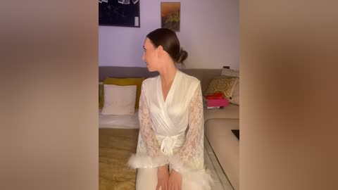 Media: Video of a woman with fair skin and dark hair in a bun, wearing a white lace robe with feather trim, sitting on a beige couch in a modern living room.