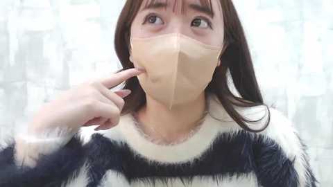 Media: Video of an Asian woman with long brown hair wearing a clear face mask and black-and-white striped sweater, holding her finger to her mouth, against a blurred cityscape background.