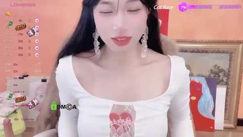 Media: A video of an Asian woman with long black hair, wearing a white off-shoulder top with a heart graphic, and dangling earrings, in a cozy, cluttered room with orange walls and art pieces.