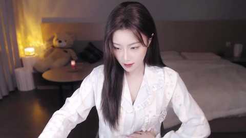 Media: Video of a young East Asian woman with long black hair, wearing a white floral kimono, sitting in a dimly lit bedroom with a bed, lamp, and stuffed teddy bear.