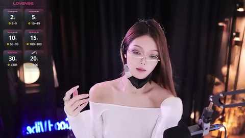 Media: Video of an Asian woman with long brown hair, wearing an off-shoulder white top and black choker, standing in a dimly lit room with a digital on-screen display and studio equipment in the background.