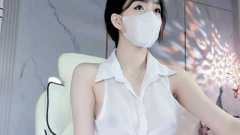 Media: Video of an East Asian woman with fair skin, wearing a white surgical mask, sleeveless white blouse, and sitting in a modern, white room with a chandelier.