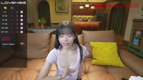 Media: A video of an Asian woman with black pigtails, wearing a purple top, sitting on a brown couch in a cozy, warmly lit living room.