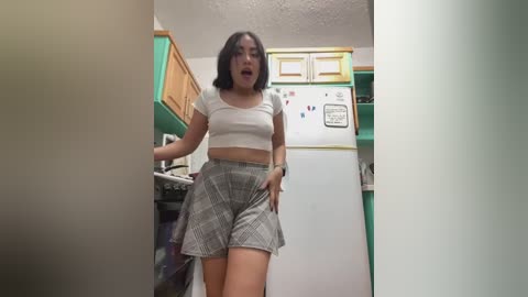 Media: Video of a young woman with shoulder-length black hair, wearing a white crop top and plaid skirt, standing in a kitchen with green cabinets, white refrigerator, and colorful magnets.