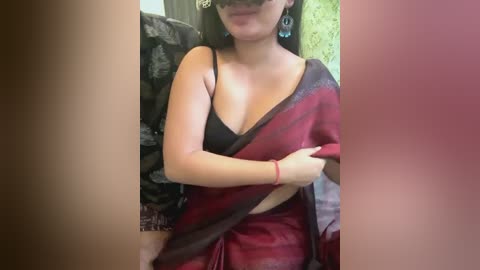 Media: Video of a woman in a red sari, revealing a black bra, wearing large blue earrings, with a blurred background of green and black patterns.