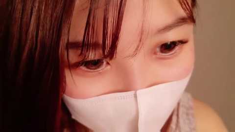 Media: Video of a young Asian woman with long, straight black hair and brown eyes, wearing a white surgical mask, looking directly at the camera.