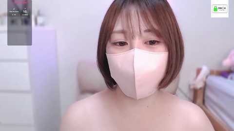 Media: Video of an Asian woman with short, straight brown hair, wearing a pink face mask, standing indoors. She has light skin and is topless. Background shows a blurred white dresser and bed.