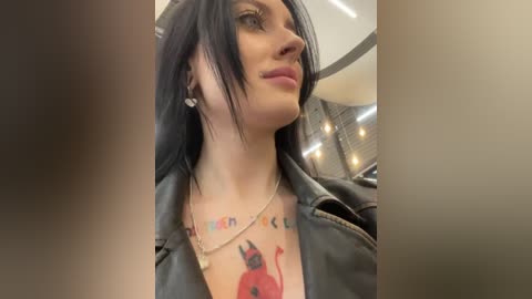 Media: Video of a woman with pale skin, long black hair, wearing a black leather jacket and necklace. She has a red devil tattoo on her chest, with a colorful \"LIVE FAST DIE YOUNG\" banner. Background shows a modern, indoor setting with a mesh wall.