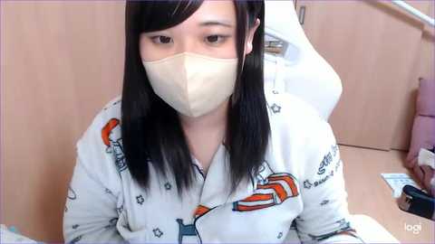 Media: Video of an East Asian woman with long black hair, wearing a white mask, pajama shirt, and headphones, seated in a wooden chair, in a home setting.