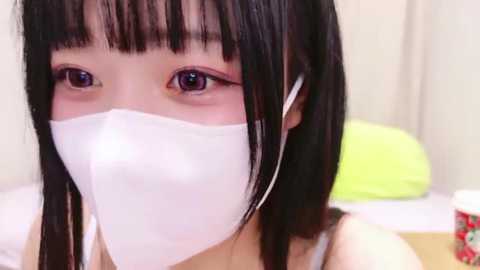 Media: Video of a young Asian woman with straight black hair, wearing a white face mask, purple eyeshadow, and white headphones, sitting on a bed with a green pillow and a red container in the background.