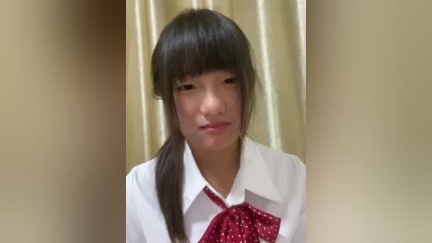 Media: Video of an East Asian girl with straight black hair, wearing a white blouse with a red polka-dot bow tie, in front of beige curtains.