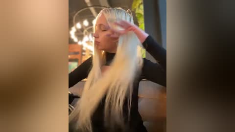 Media: Video of a blonde woman with long, straight hair in a black top, adjusting her hair in a modern, well-lit room with a chandelier and plants in the background.