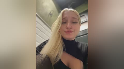 Media: Video of a young blonde woman with fair skin, wearing a black turtleneck, making a pouty face in a dimly lit, tiled bathroom with green walls.