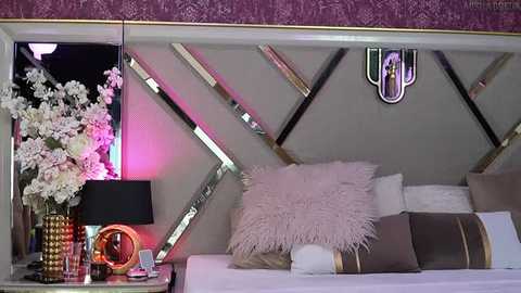 Media: Video of a stylish bedroom with a modern design featuring a white bed, gold-framed mirrors, white fluffy pillow, and a purple wall. A black lamp with a pink shade sits on a nightstand adorned with a gold orb.