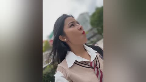 Media: Video of a young Asian woman with medium skin tone, wearing a white shirt and beige cardigan, red tie, and long black hair, standing outdoors with blurred greenery and buildings in the background.