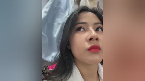 Media: Video of a young woman with medium skin tone, long dark hair, and bright red lipstick, looking contemplative. Background features a white shirt and blurred blue fabric.