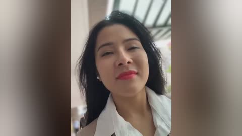 Media: Video of a smiling East Asian woman with long black hair, wearing a white shirt, against a blurred background featuring greenery and a roof.