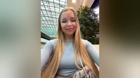 Media: Video of a smiling young woman with long blonde hair, wearing a light blue long-sleeve top, sitting in a modern indoor space with glass walls and green plants.