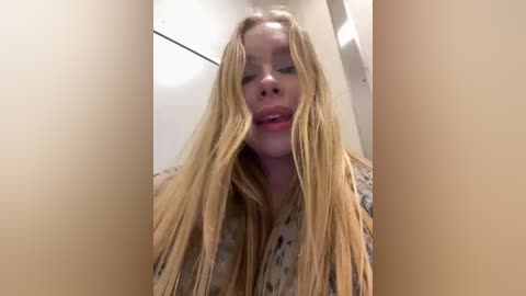 Media: A video of a young woman with long, wet blonde hair covering her face, wearing a patterned top, standing indoors against a plain white wall.