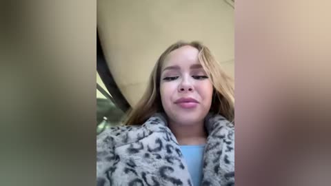 Media: A video of a young Caucasian woman with long blonde hair, wearing a gray leopard-print coat, lying down in a beige interior. The image is slightly blurred.