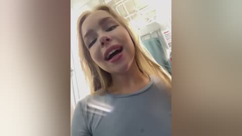 Media: A video of a young blonde woman with fair skin and closed eyes, wearing a light blue shirt, in a blurry bathroom setting.