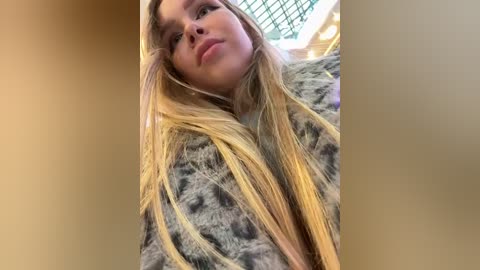 Media: Video of a young woman with long, straight blonde hair, wearing a grey fur coat, looking slightly to the side with a neutral expression, in an indoor setting with a green metal ceiling.