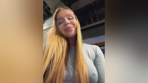 Media: Video of a young, blonde woman with long hair, wearing a light gray top, smiling with eyes closed, taken indoors with dim lighting.