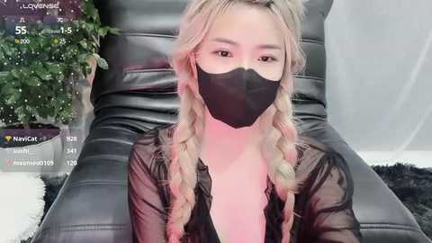 Media: Video of an Asian woman with long blonde hair, wearing a black face mask, black top, and sitting on a black leather couch. The background includes a green plant and a digital screen displaying temperature and humidity data.