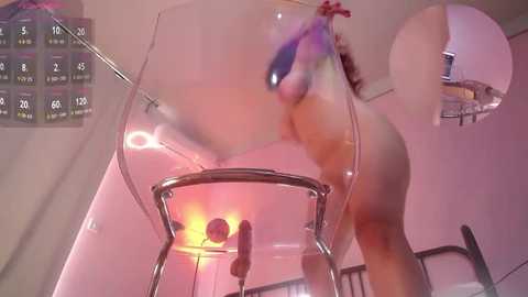 Media: A blurred, motion-blurred video of a naked woman with medium-sized breasts, wearing a pink headband, bending over a chair in a pink room with a wall-mounted calendar and visible ceiling light.