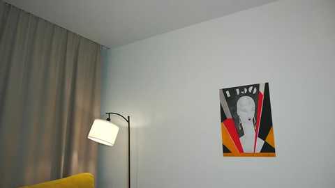 Media: A minimalist room with a white wall featuring a modern, abstract painting of a face with bold red, black, and yellow lines. A yellow armchair and a floor lamp with a white shade are visible in the background.