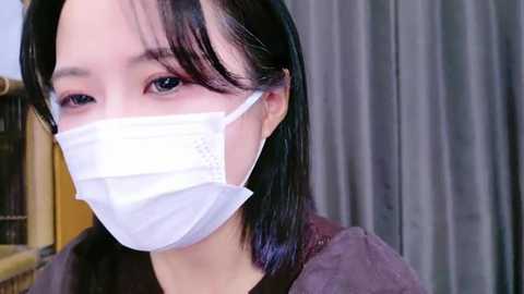 Media: Video of an East Asian woman with straight black hair, wearing a white medical mask, grey shirt, and grey curtains in the background.