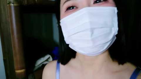 Media: A close-up video of an East Asian woman with straight, black hair, wearing a white surgical mask, light blue spaghetti strap top, and a wooden shelf background.