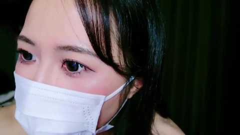 Media: Video of an Asian woman with straight black hair, wearing a white surgical mask, and dark eyes, in a dimly lit room.