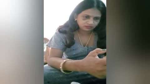 Media: Video of a South Asian woman with long black hair and a medium skin tone, wearing a grey T-shirt and jeans, sitting in a wooden chair, holding a phone, with a blurred background.