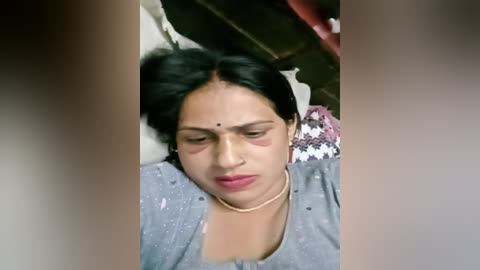 Media: Video of a woman lying on a bed, wearing a gray shirt, eyes closed, with a tear on her cheek, surrounded by blurred, indistinct figures.