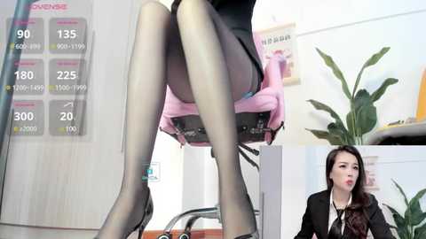 Media: Video of a woman in a black suit, seated on a pink office chair, wearing sheer black stockings and black heels. She has long, dark hair. Background includes a white wall with a calendar, green plant, and desk.