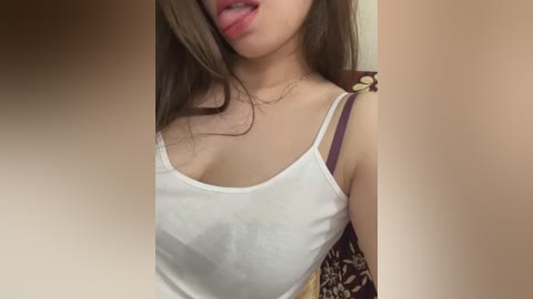 Media: Video of an Asian woman with fair skin, long brown hair, wearing a white spaghetti-strap tank top, sticking out her tongue, in a beige room.