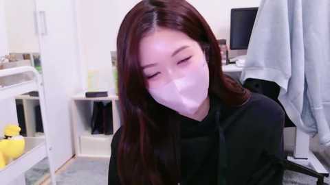 Media: Video of an East Asian woman with long, straight brown hair, wearing a black dress and a white mask, sitting in a clean, white-tiled room with shelves, a computer, and a hanging jacket.