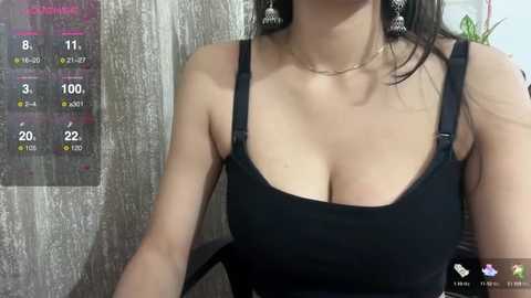 Media: Video of a light-skinned woman with long dark hair, wearing a black tank top that accentuates her large breasts, standing against a textured wooden wall.