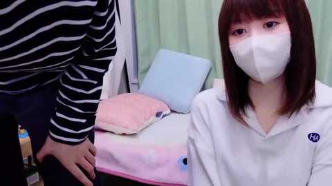 Media: Video of a young Asian woman with straight brown hair, wearing a white mask and white hoodie, sitting on a bed with a blue pillow and pink blanket, next to a person in a black-and-white striped shirt.