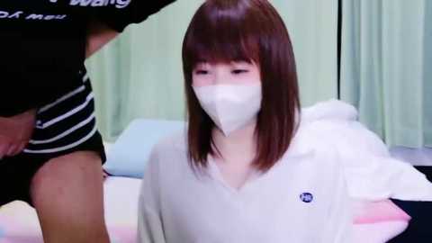 Media: Video of a young East Asian woman with straight brown hair, wearing a white mask and hoodie, sitting on a bed in a clinical room with green curtains and a striped man standing nearby.