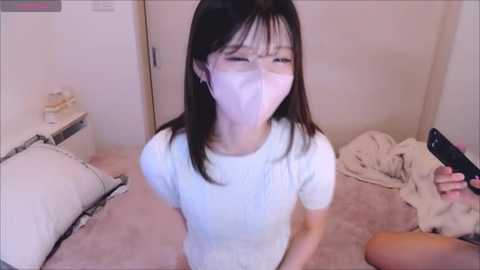 Media: Video of an East Asian woman in a white mask, white top, and glasses, standing in a cluttered bedroom with unmade bed, beige carpet, and a phone in hand.