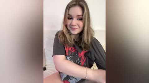 Media: A video of a young Caucasian woman with shoulder-length brown hair, wearing a dark graphic t-shirt, sitting on a bed with white sheets. The background is blurred.