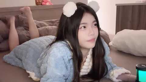 Media: Video of a young Asian woman with long black hair, wearing bear ears and a light blue fuzzy robe, lying on a bed, looking contemplative.