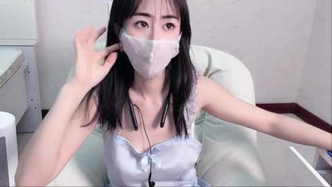 Media: Video of an East Asian woman with long black hair, wearing a light blue satin camisole and white surgical mask, seated in a hospital bed. Background features a white wall and medical equipment.