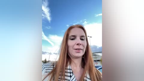 Media: Video of a fair-skinned, red-haired woman wearing a black-and-white checkered jacket, standing outdoors with a clear blue sky and white clouds in the background.