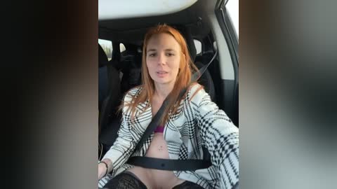 Media: Video of a young woman with light skin, long red hair, wearing a black and white plaid jacket, sitting in a car with a seatbelt on, looking slightly surprised.