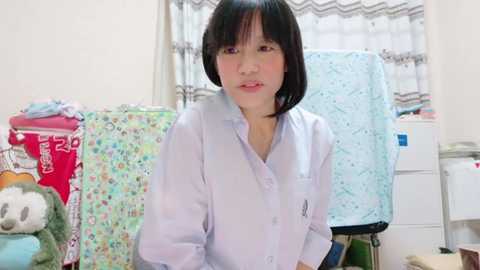 Media: A video of an East Asian woman with short black hair, wearing a light blue shirt, standing in a cluttered room with colorful blankets and stuffed animals.