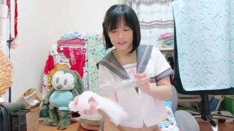 Media: Video of an Asian woman with short black hair, wearing a sailor-style school uniform, examining a plush toy in a cluttered, brightly lit room filled with clothing and bags.