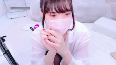 Media: Video of a young East Asian woman in a white lab coat, face mask, and gloves, kneeling in a sterile lab setting, holding her hands together, looking thoughtful.